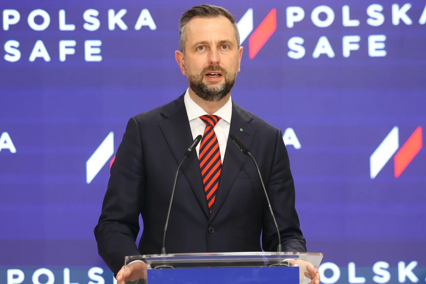Wicepremier i szef MON Władysław Kosiniak-Kamysz Władysław Kosiniak-Kamysz przy mównicy, w garniturze i czerwono-czarnej krawacie, na tle ścianki POLSKA SAFE.