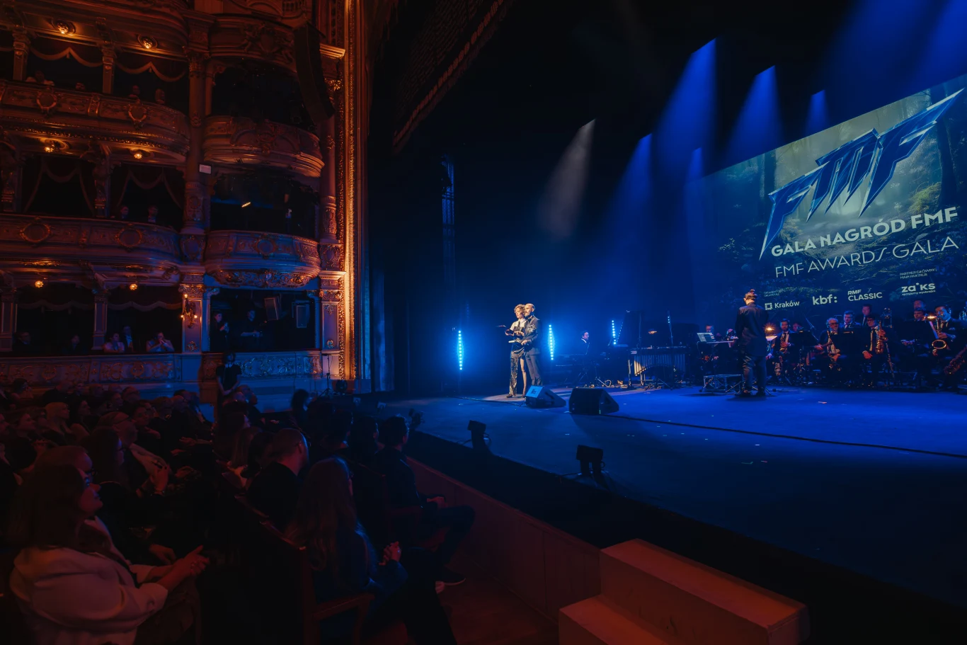 Ludzka sylwetka odbierająca nagrodę na oświetlonej scenie podczas gali FMF, w otoczeniu muzyków orkiestrowych i eleganckiej widowni siedzącej w klasycznej sali teatralnej.