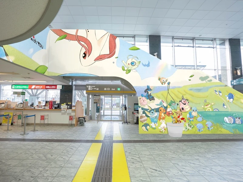 Kolorowa ściana ozdobiona dużym muralem z postaciami Pokémon, nowoczesne wnętrze budynku z szerokim wejściem i jasnymi, przeszklonymi ścianami, wyraźnie widoczne ścieżki dla osób niedowidzących wyznaczone żółtym pasem na podłodze.