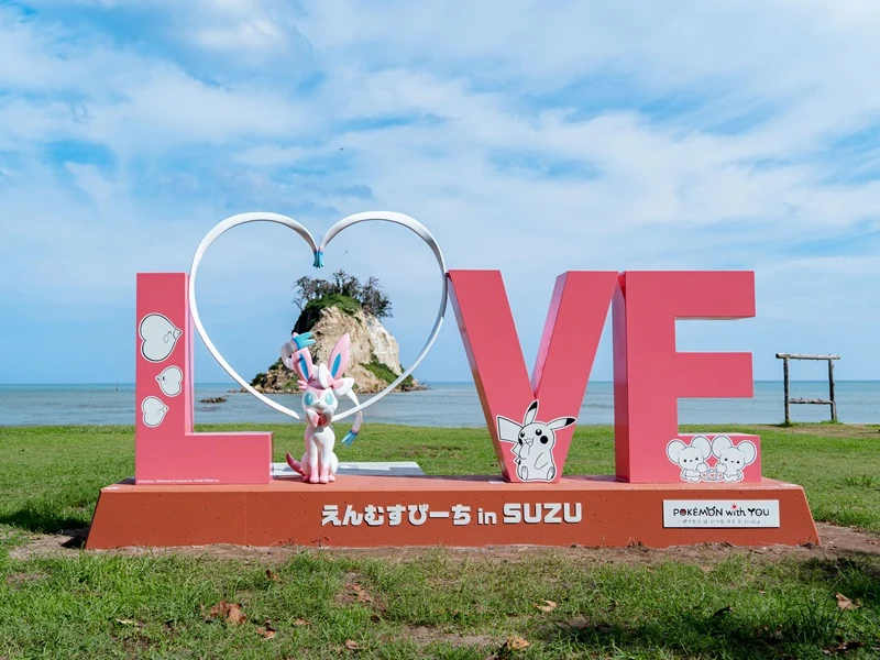 Różowy napis LOVE z postacią Sylveona i motywami Pokémon, ustawiony na tle wyspy pokrytej zielenią pośrodku morza pod niebieskim niebem.