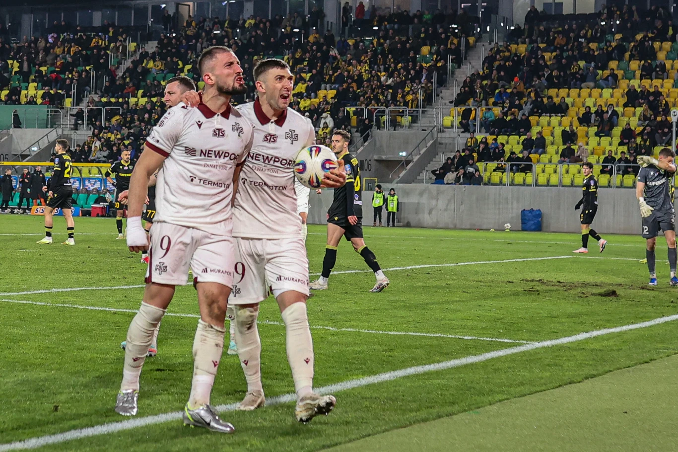 Widzew Łódź walczy o utrzymanie Dwóch piłkarzy w jasnych strojach celebruje zdobycie gola, trzymając piłkę i okazując radość, w tle widoczna trybuna pełna kibiców.