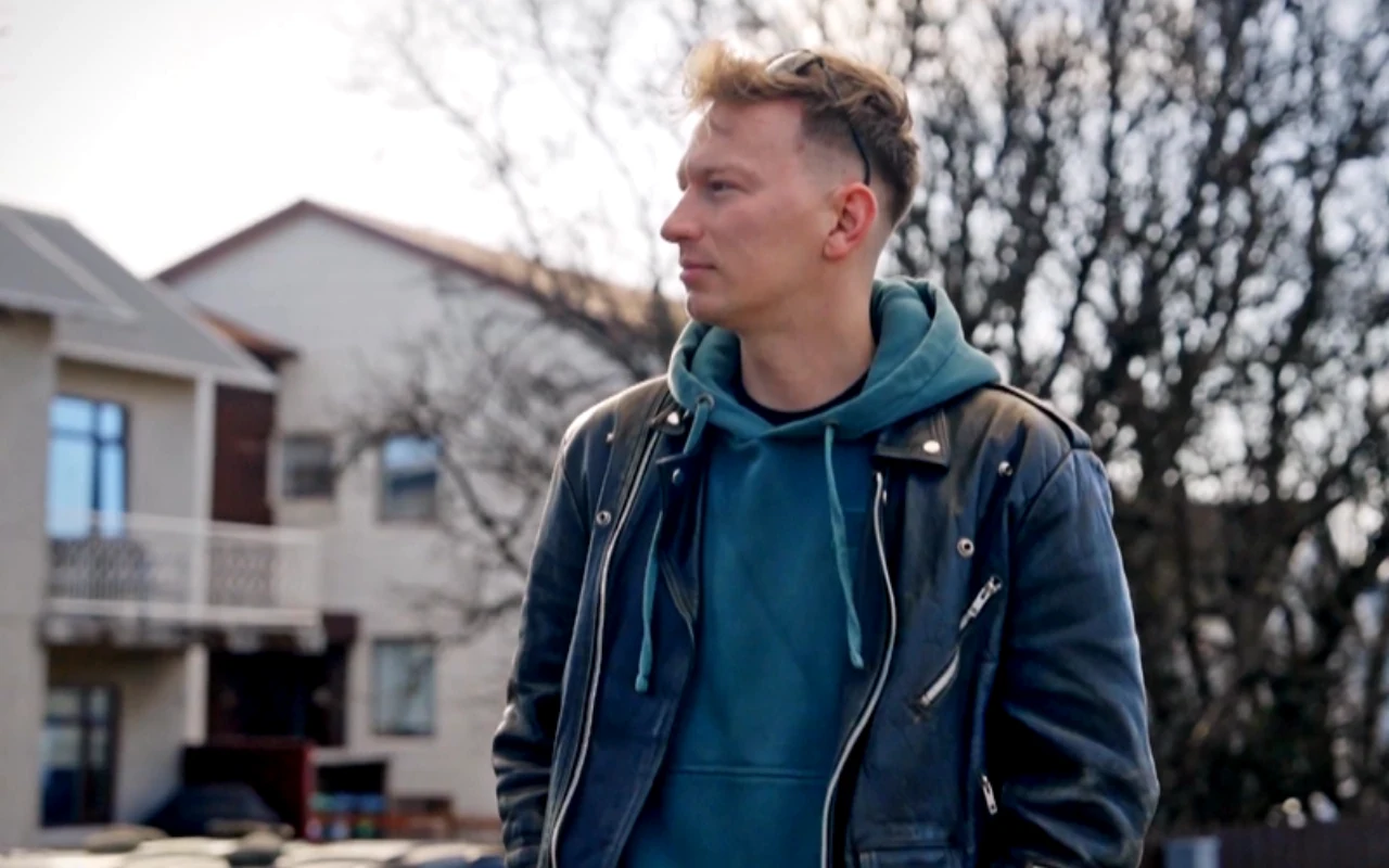 Mężczyzna w skórzanej kurtce i niebieskiej bluzie z kapturem stoi na zewnątrz, w tle widoczne jasne budynki i bezlistne drzewo.