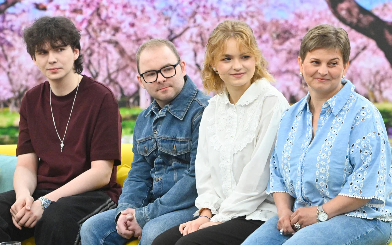 Marcycha z najbliższymi w studiu "halo tu polsat" Czwórka osób siedzących na żółtej sofie, uśmiechnięci, ubrani w codzienne, wygodne stroje, na tle wiosennych drzew kwitnących na różowo.