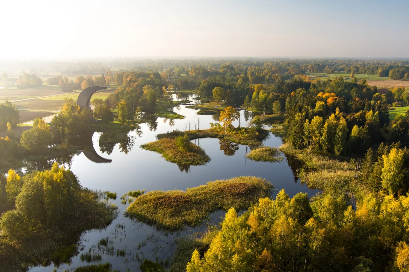 Bagna i torfowiska w naturalny sposób utrudniają operacje militarne Jezioro otoczone gęstym lasem mieszanym z dominacją drzew liściastych w jesiennych barwach, nad wodą unosi się lekka mgła, a w tle rozciągają się pola uprawne.