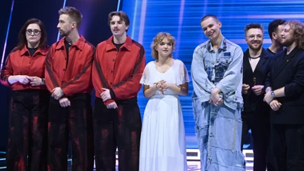 Grupa osób stojących na scenie, część ubrana w czerwone koszule i czarne spodnie, kobieta w białej sukience oraz mężczyzna w jasnoniebieskim, jeansowym komplecie; w tle podświetlone niebieskim światłem pionowe pasy.