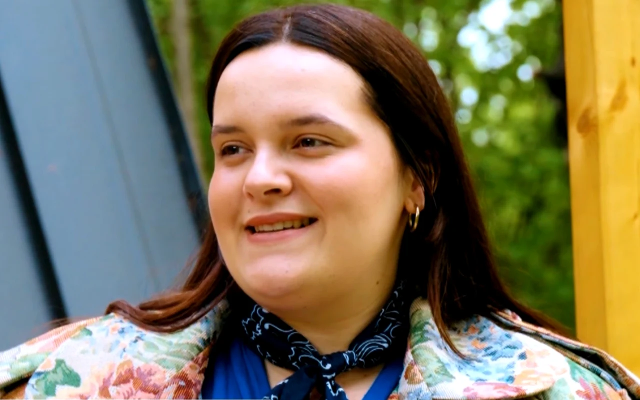 Uśmiechnięta młoda kobieta z długimi brązowymi włosami, ubrana w kolorową kurtkę i granatową chustkę na szyi, stojąca na tle zieleni i drewnianej konstrukcji.