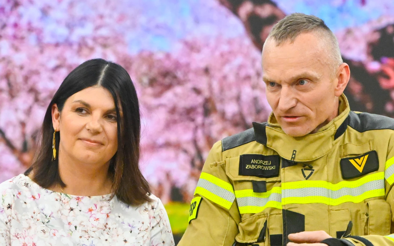 Kobieta w jasnej, kwiecistej bluzce stoi obok mężczyzny w stroju strażaka z nazwiskiem na mundurze, oboje patrzą w tym samym kierunku.