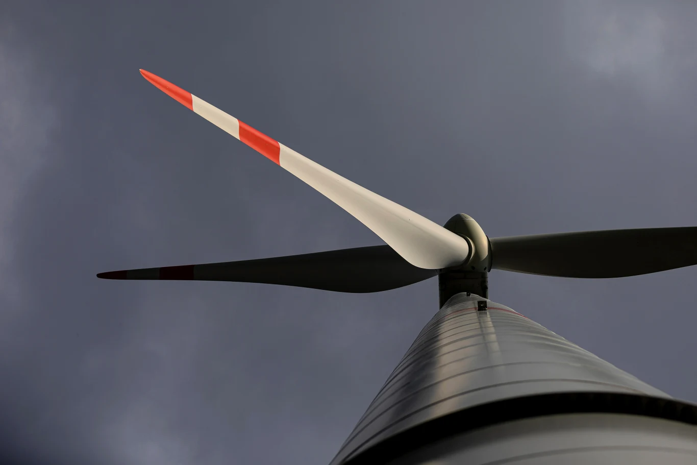 Nowoczesna turbina wiatrowa o trzech łopatach, zbliżenie na wysoką wieżę oraz śmigła z czerwonymi końcówkami, ciemne, burzowe chmury na tle nieba.