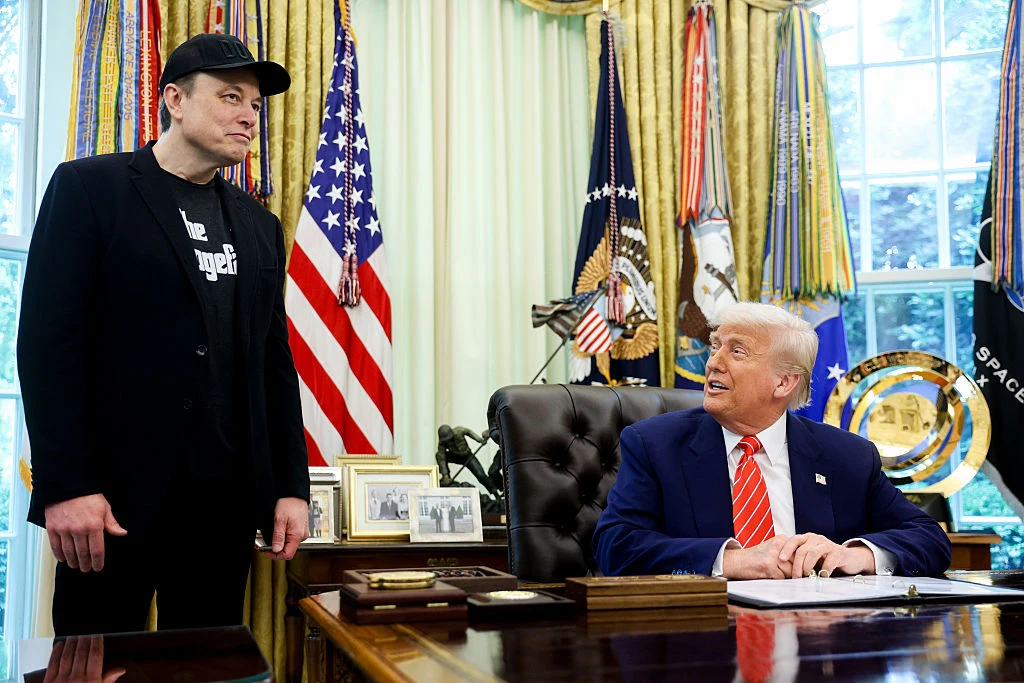 Elon Musk i Donald Trump Dwóch mężczyzn znajduje się w gabinecie o wystroju prezydenckim; jeden siedzi przy dużym biurku w garniturze i czerwonym krawacie, drugi stoi obok w casualowym stroju z czapką z daszkiem, między nimi widoczna jest interakcja wzrokowa.