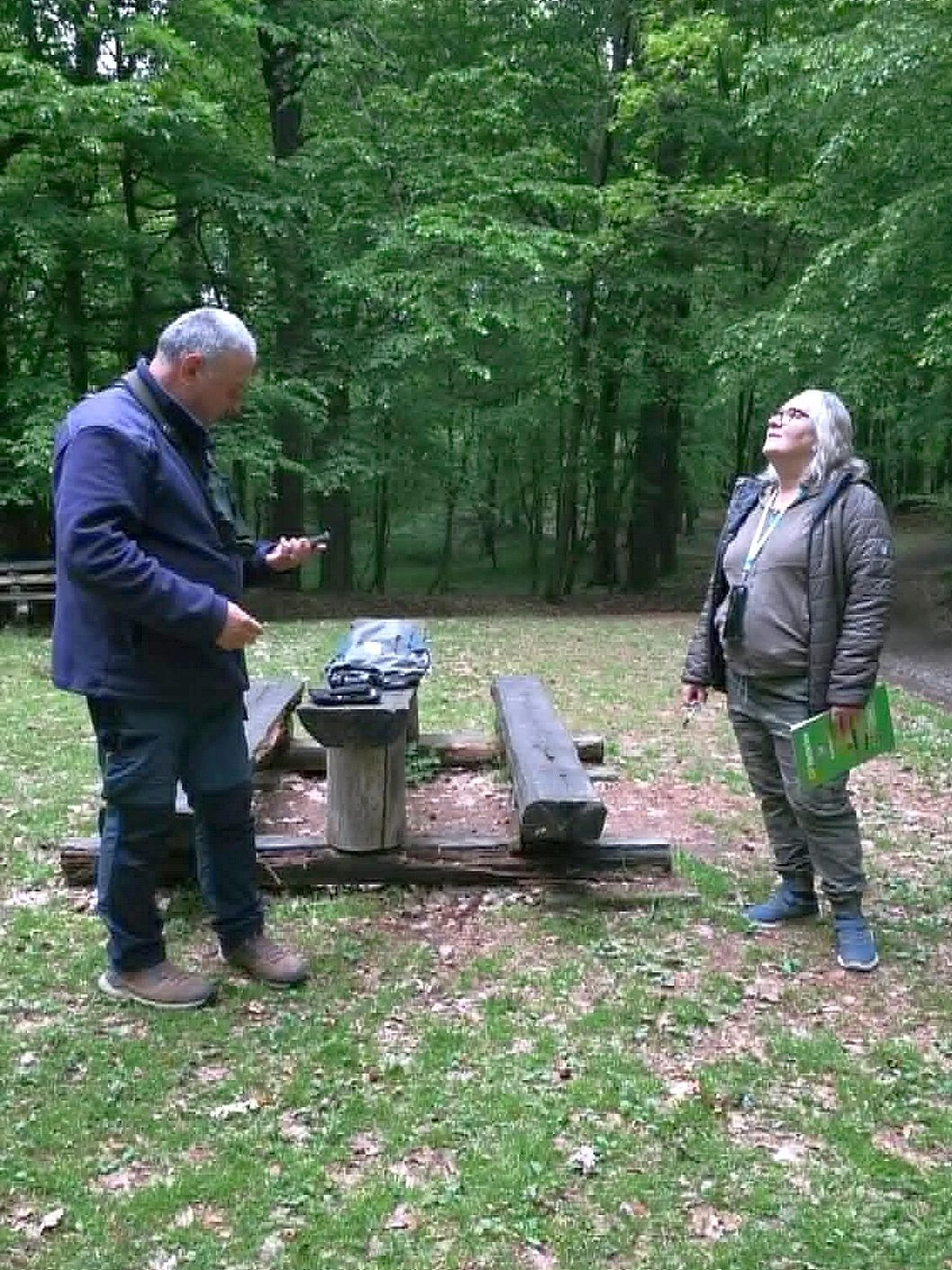 Rajdowe nasłuchiwanie ptaków w lesie o poranku Dwie osoby stoją przy drewnianym stole piknikowym wśród drzew, jedna trzyma otwarty zeszyt, druga telefon, na stole leży torba oraz mapa.