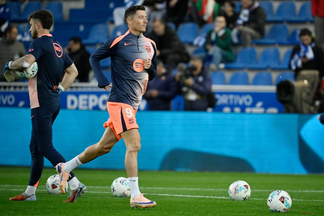 Piłkarz w stroju treningowym FC Barcelony biegnie po murawie wśród kilku piłek podczas rozgrzewki na stadionie.