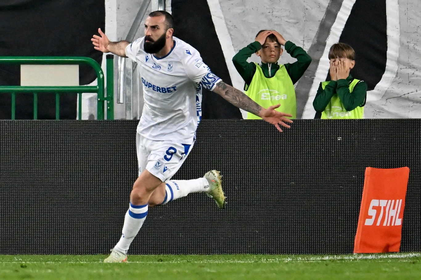 Mikael Ishak w meczu z Radomiakiem Piłkarz drużyny w białych strojach świętuje zdobycie gola z rozłożonymi rękami, w tle dwaj młodzi chłopcy w zielonych kurtkach wyrażają zaskoczenie i rozczarowanie.