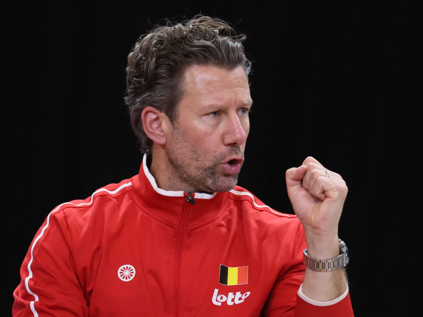 Wim Fissette Mężczyzna w czerwonej sportowej kurtce z logo oraz flagą Belgii, zaciśnięta pięść uniesiona w geście determinacji, na czarnym tle.