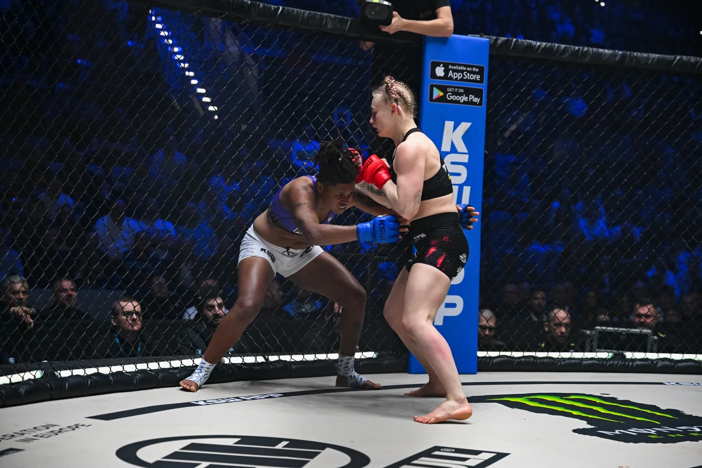 Dwie zawodniczki MMA walczą w oktagonie, jedna w czarnych spodenkach i czerwonych rękawicach dociska rywalkę w białych spodenkach do siatki.