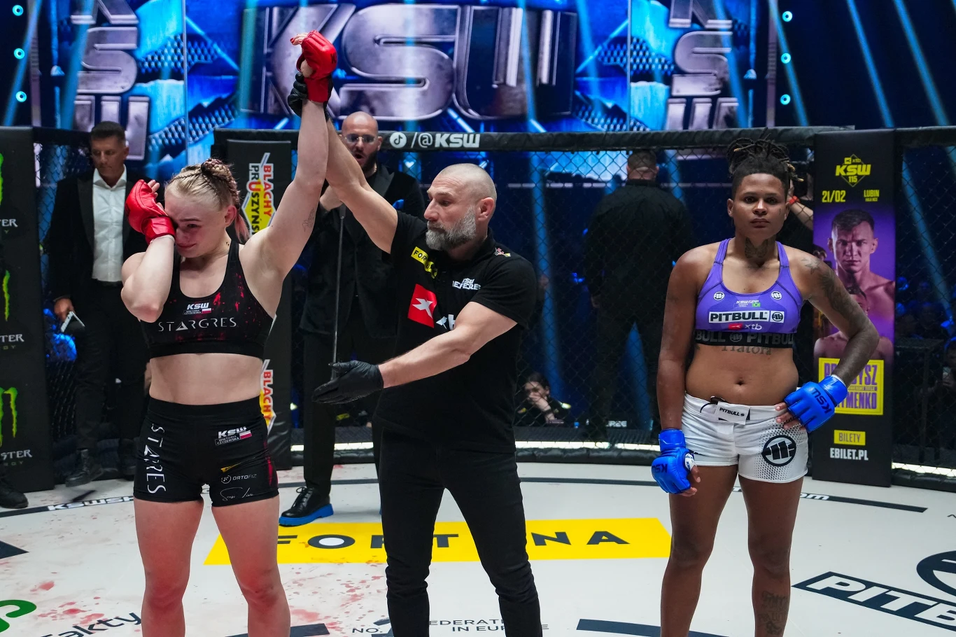 Sędzia unosi rękę zwyciężczyni walki MMA, podczas gdy druga zawodniczka stoi obok, wyrażając rozczarowanie wynikiem.