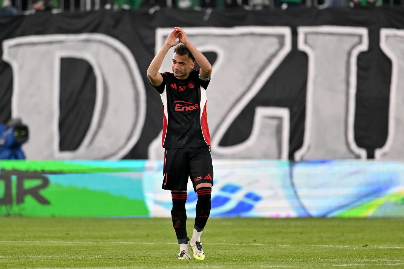 Leandro żegna się z kibicami Radomiaka Piłkarz w czarno-czerwonym stroju wykonuje gest uniesionych rąk, stojąc na murawie stadionu na tle dużego napisu na trybunach.