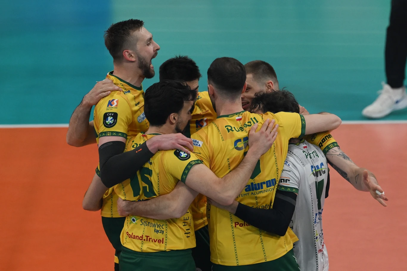 Aluron CMC Warta Zawiercie Grupa siatkarzy w żółtych koszulkach świętuje razem, obejmując się na boisku sportowym po udanej akcji.