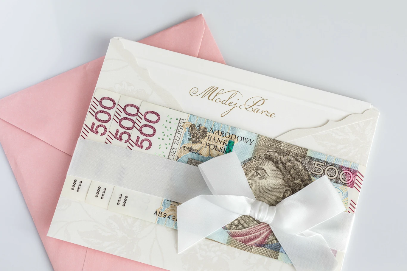 Co trzeci Polak rezygnuje z udziału w weselu z powodu związanych z tym wysokich kosztów Biała koperta z napisem "Młodej Parze", biała wstążka, banknoty 500 zł, różowa koperta w tle.