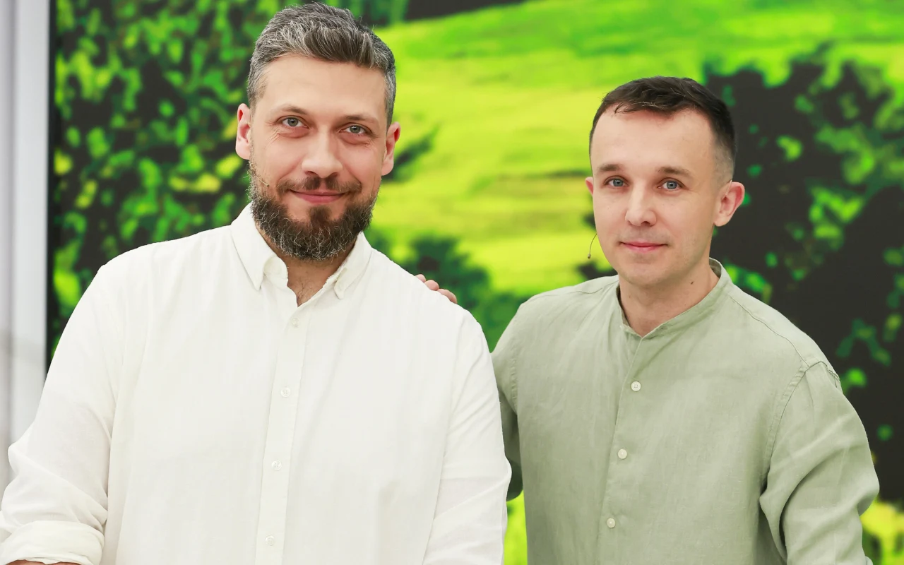 Dwóch mężczyzn uśmiechających się do kamery, ubrani w jasne koszule, na tle rozmytego zielonego krajobrazu.