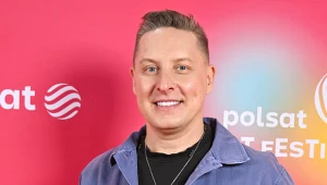 Mężczyzna w jasnoniebieskiej kurtce i czarnym t-shircie na tle czerwonej ścianki z logotypami Polsat i Festiwalu.