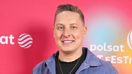 Mężczyzna w jasnoniebieskiej kurtce i czarnym t-shircie na tle czerwonej ścianki z logotypami Polsat i Festiwalu.