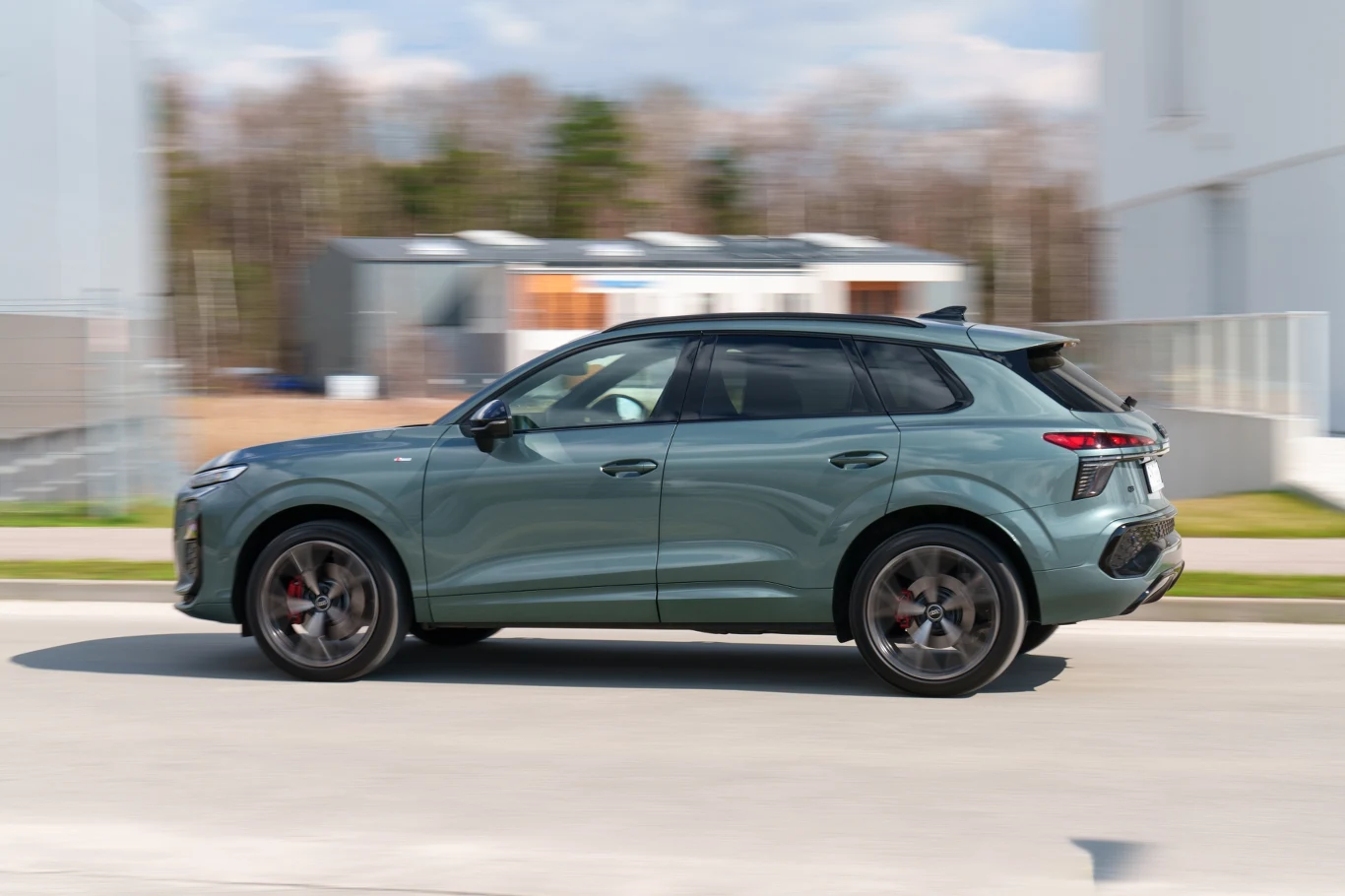 Zielony SUV poruszający się po asfaltowej drodze, z nowoczesną linią nadwozia i przyciemnianymi szybami, na tle niskich, jasnych budynków.
