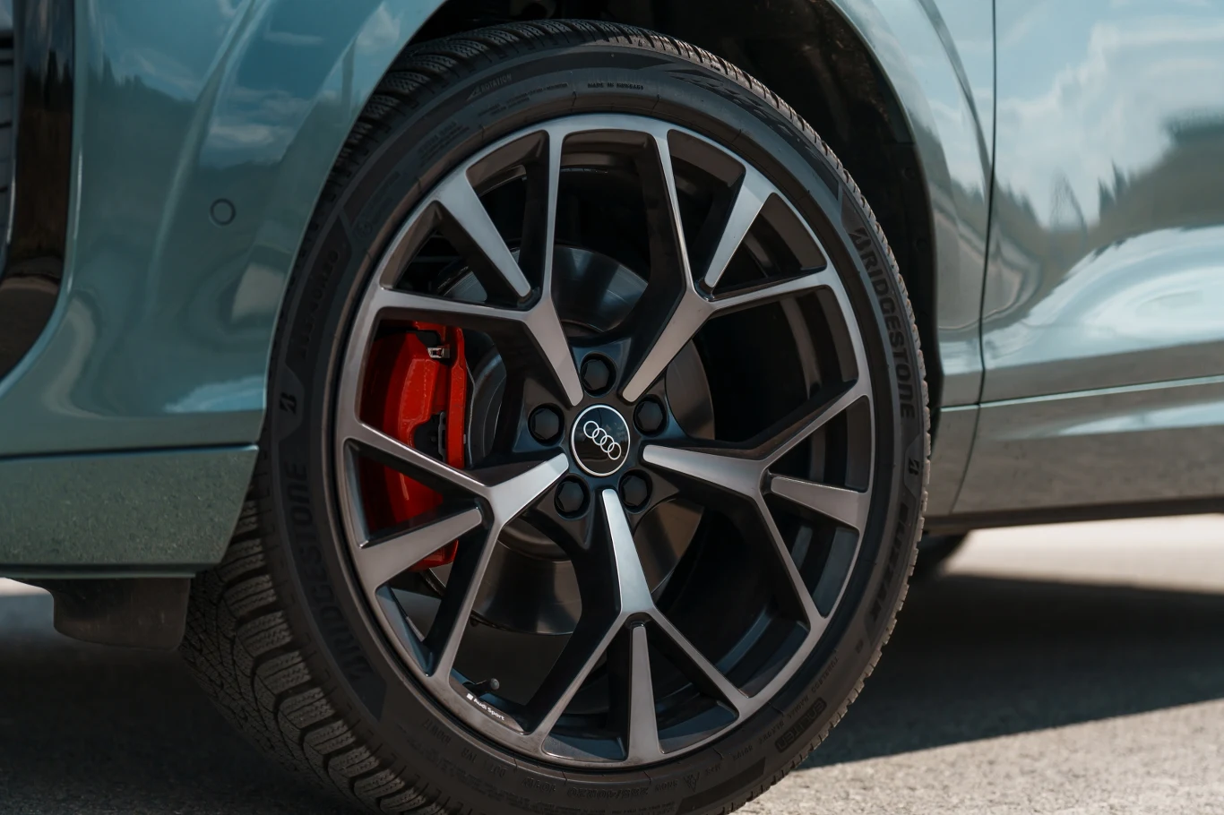 Srebrna felga aluminiowa Audi z widocznym czerwonym zaciskiem hamulcowym na tle jasnoniebieskiego nadwozia auta.