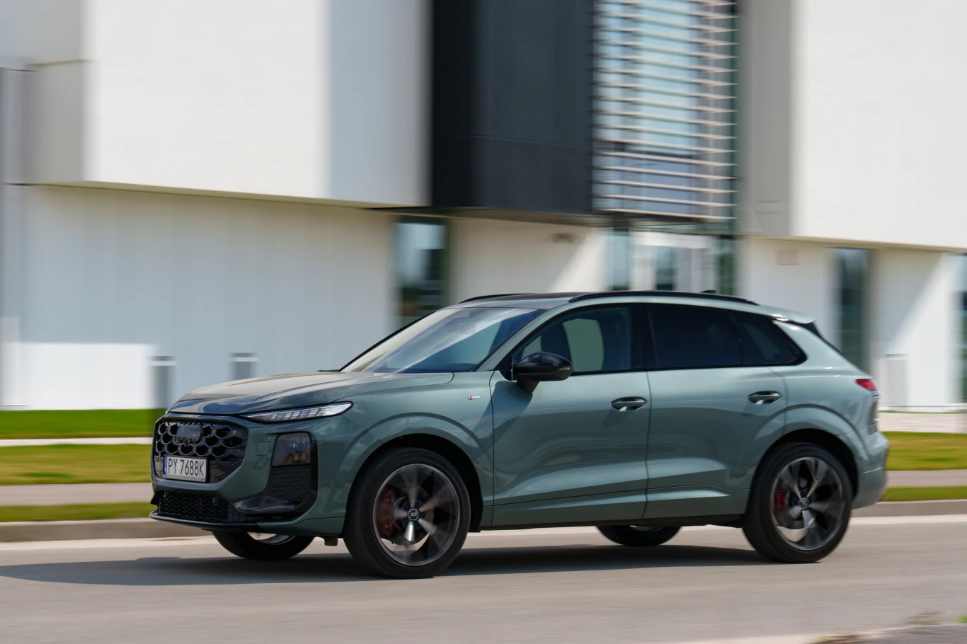 Jasnozielony SUV Audi Q4 e-tron w ruchu na miejskiej ulicy, schludnie zaparkowane przy nowoczesnym, białym budynku z dużymi oknami.