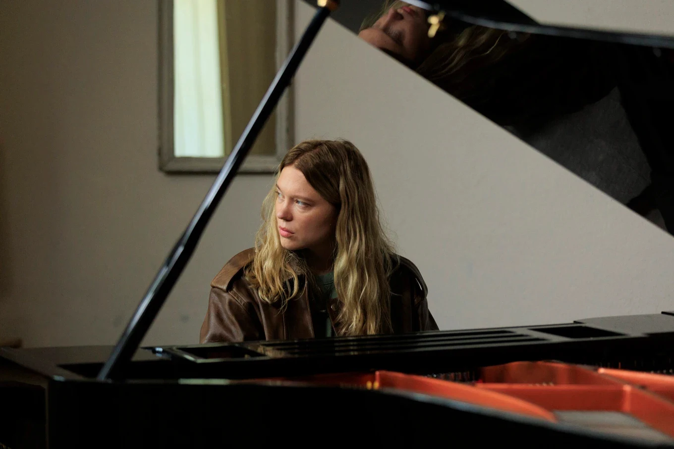 Léa Seydoux w filmie Marie Kreutzer “Gentle Monster” Léa Seydoux jako młoda kobieta z długimi blond włosami, siedzi przy fortepianie w brązowej kurtce, patrzy w bok.