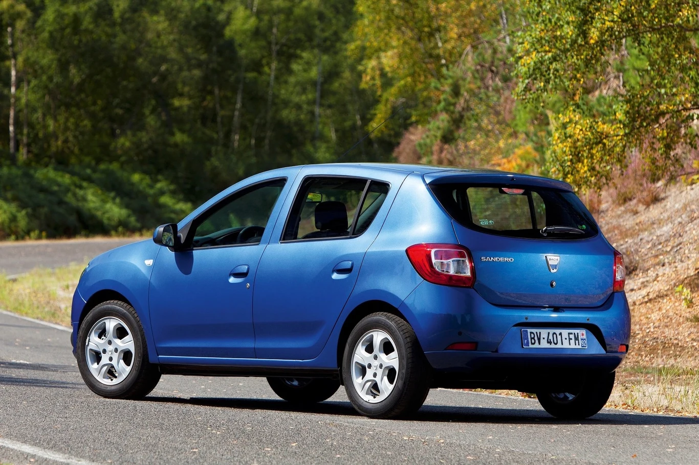 Niebieska Dacia Sandero zaparkowana przy leśnej drodze, otoczona zielonymi drzewami.