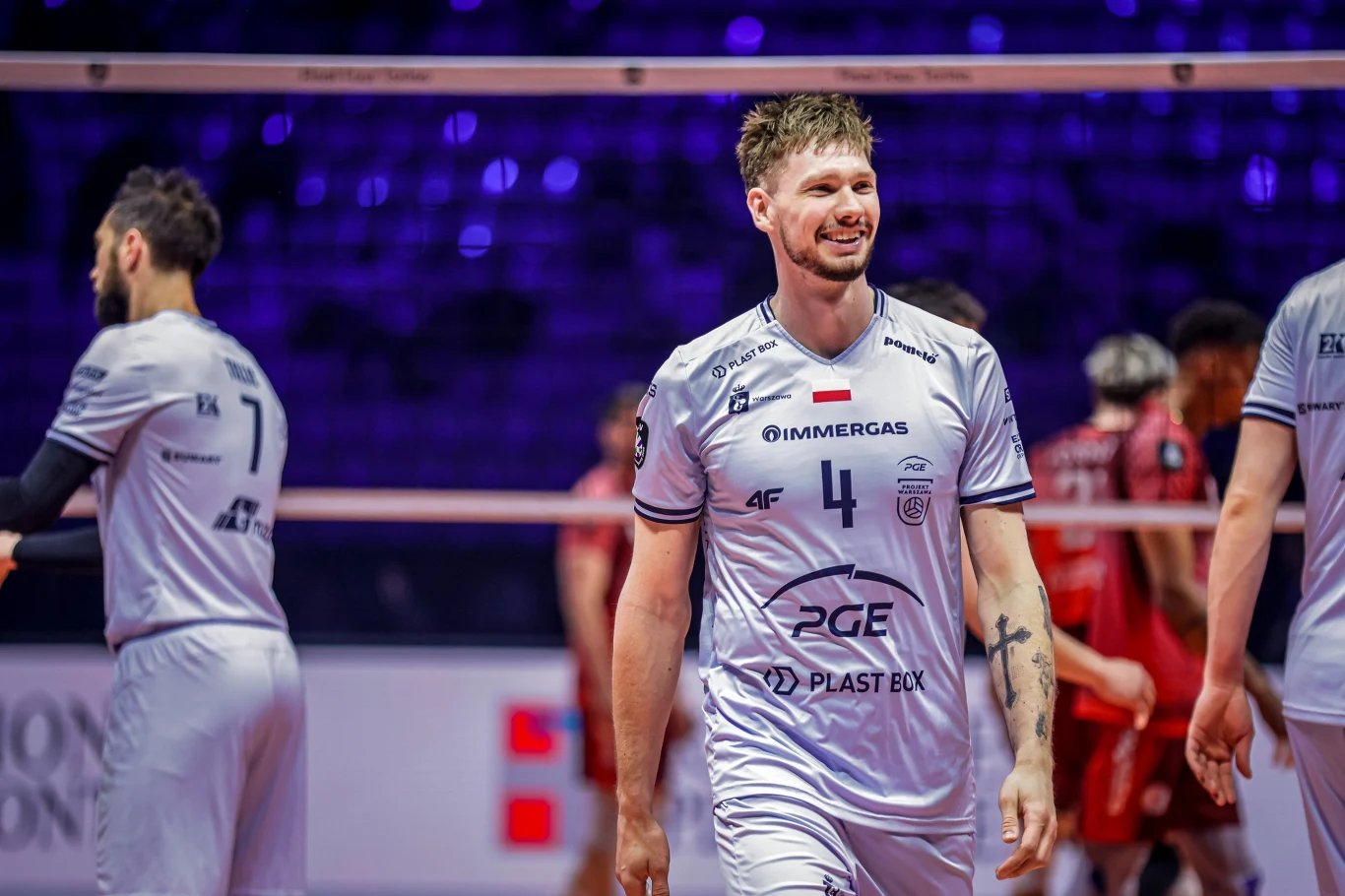 Jakub Kochanowski Uśmiechnięty siatkarz w białym stroju z numerem 4 na piersiach wychodzi na boisko, w tle widoczni pozostali zawodnicy oraz siatka.