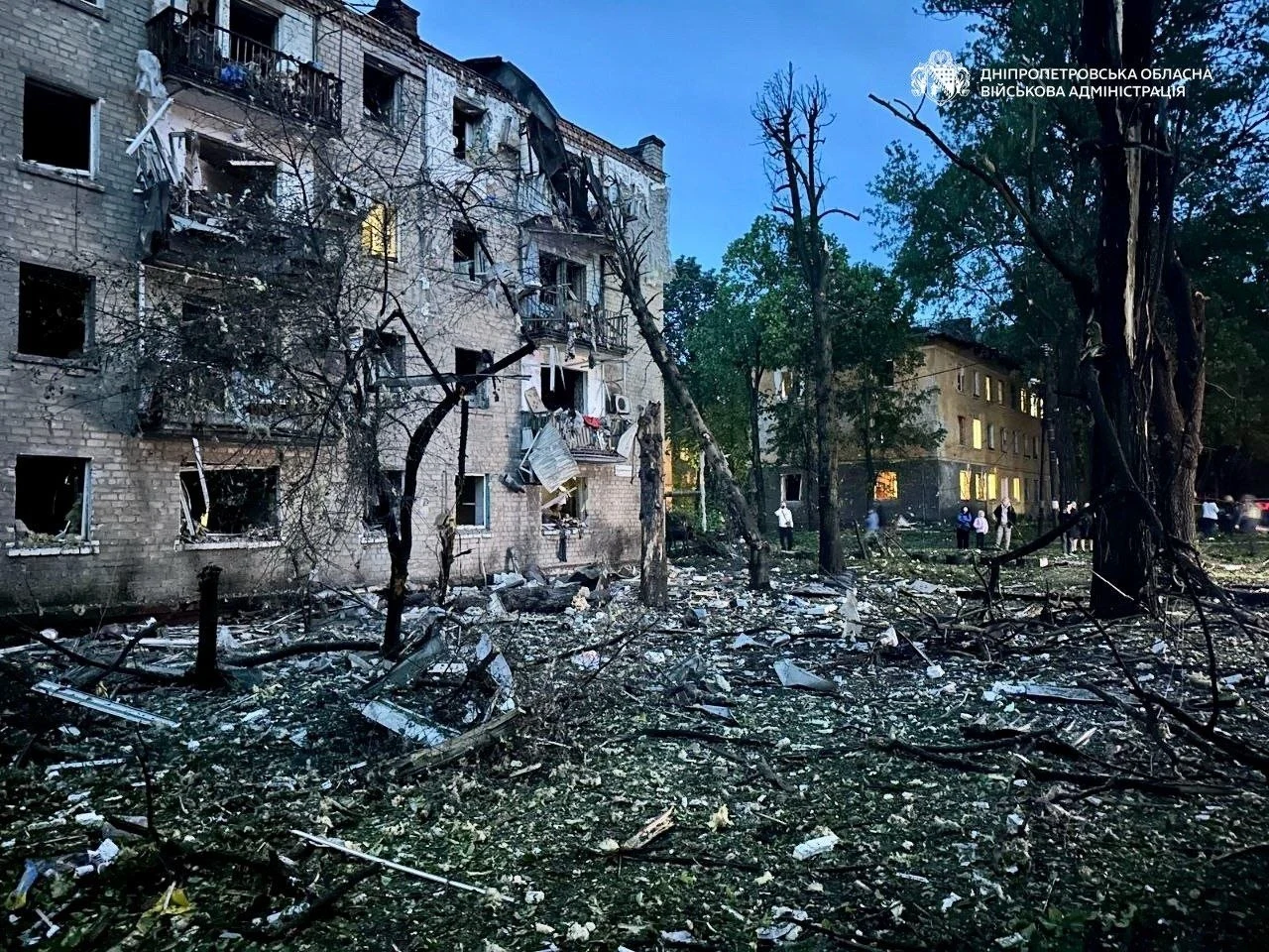 Uszkodzony blok mieszkalny z licznymi wybitymi oknami, zniszczoną elewacją i resztkami po eksplozji; wokół rozrzucone gruzy, połamane drzewa i poraniona zieleń, w oddali widoczne inne budynki.