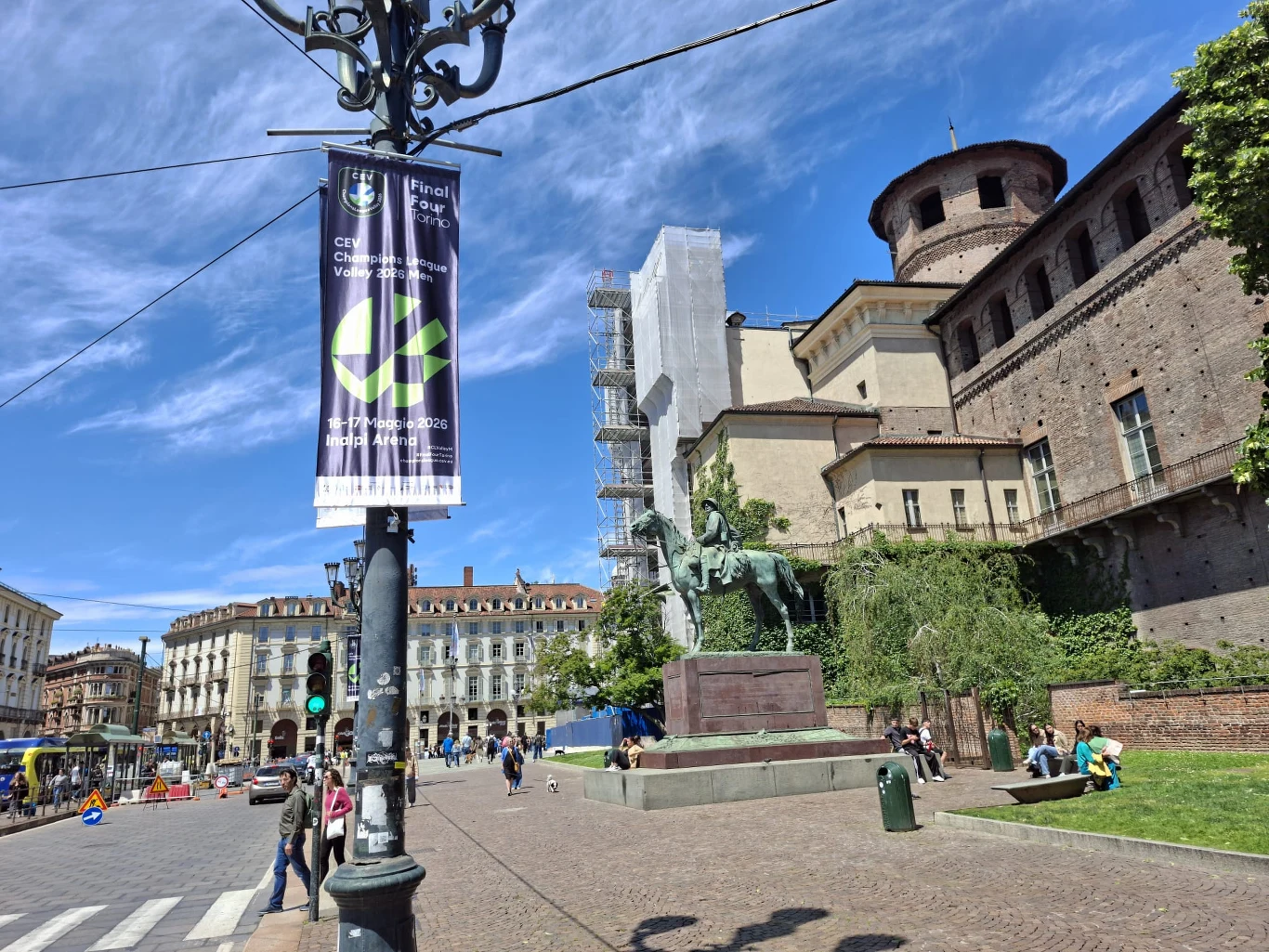 Jeden z banerów, a w zasadzie proporczyków reklamujących turniej, w pobliżu Piazza Castello w Turynie Pomnik jeźdźca na koniu przed zabytkowym budynkiem z wieżą, zielone drzewa i ludzie odpoczywający na chodniku w słoneczną pogodę.