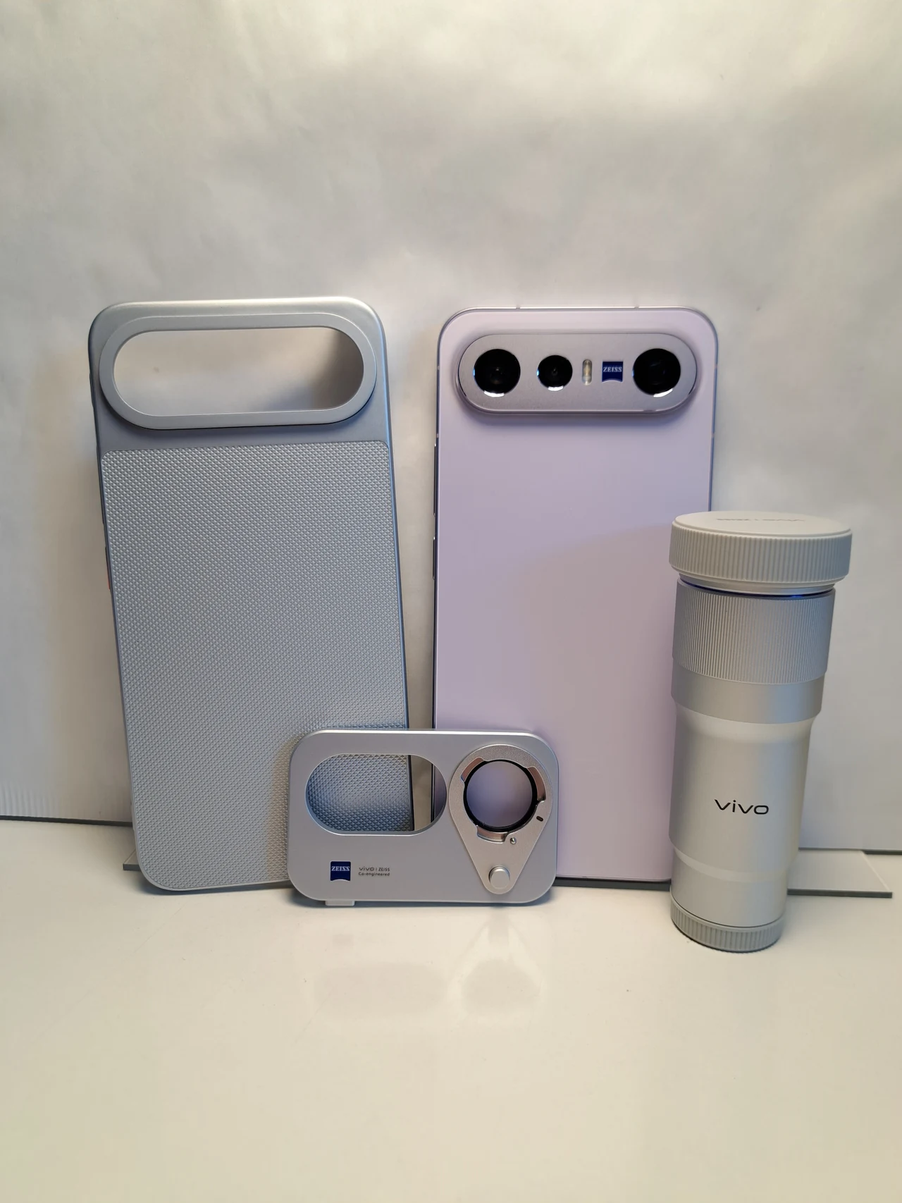Cztery akcesoria elektroniczne w jasnych barwach: smartfon z trzema aparatami na tylnej obudowie, duży głośnik, obiektyw do telefonu oraz nakładka z lupą, wszystkie sygnowane logiem Zeiss lub Vivo.