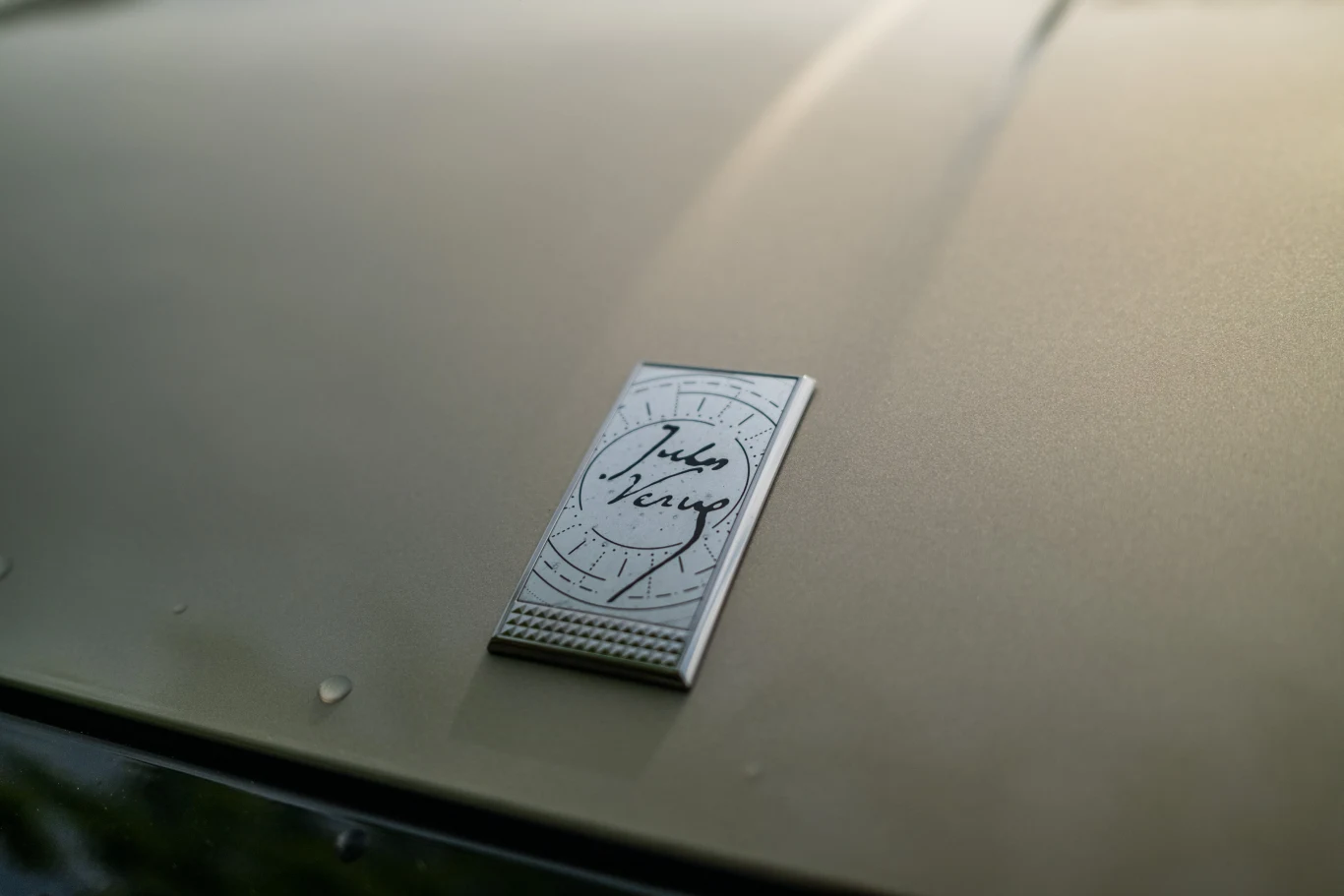 Metalowa tabliczka z podpisem umieszczona na gładkiej, jasnozłotej powierzchni maski samochodu.