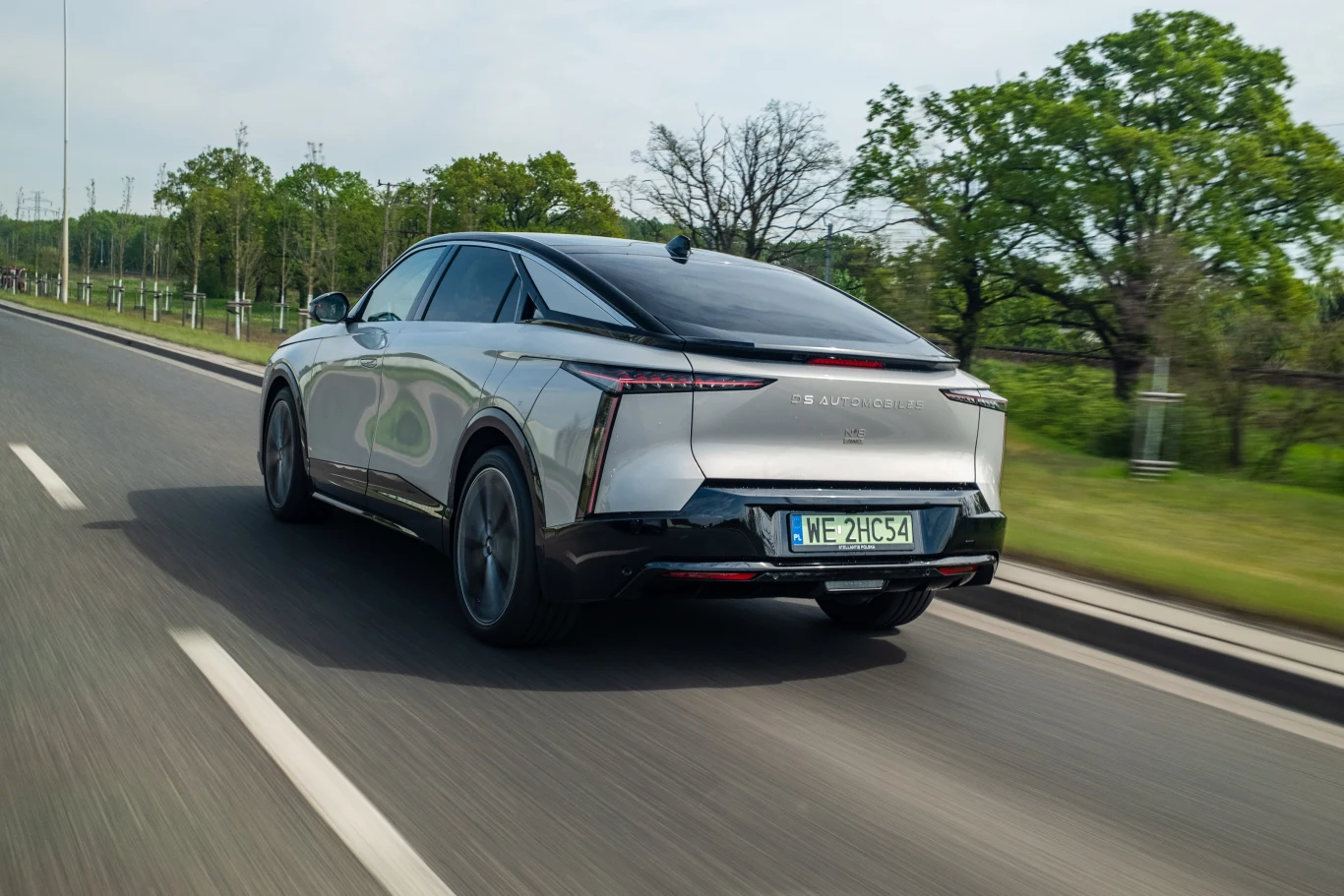 Nowoczesny, srebrny samochód elektryczny o futurystycznym designie porusza się dynamicznie po asfaltowej drodze otoczonej zielenią i drzewami.