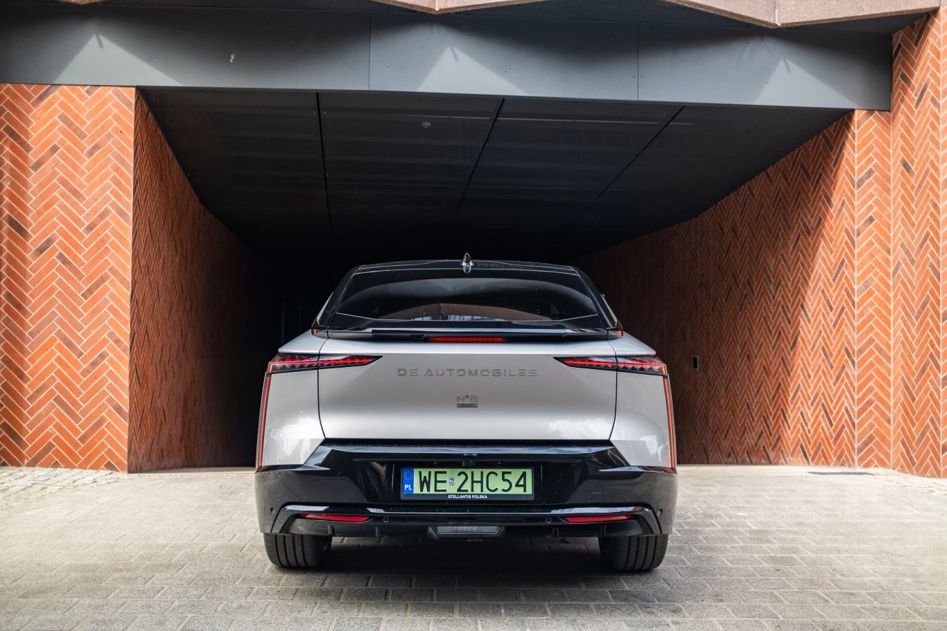 Srebrny samochód DS Automobiles zaparkowany tyłem przed wjazdem do garażu o nowoczesnej, ceglanej elewacji.