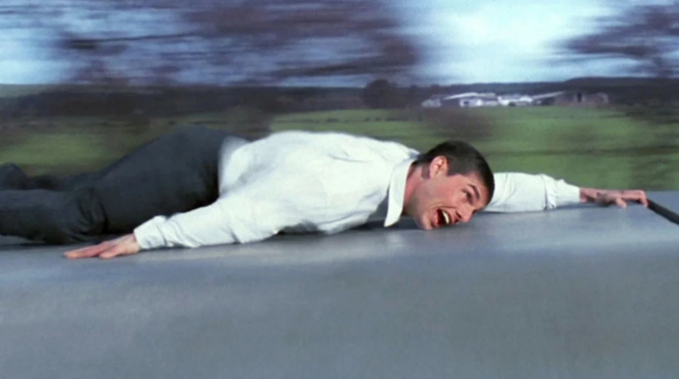 Tom Cruise w finale filmu "Mission: Impossible" Mężczyzna w białej koszuli leży płasko na dachu samochodu i trzyma się kurczowo, z wyrazem przerażenia na twarzy, w tle rozmazane drzewa wskazujące na dużą prędkość pojazdu.