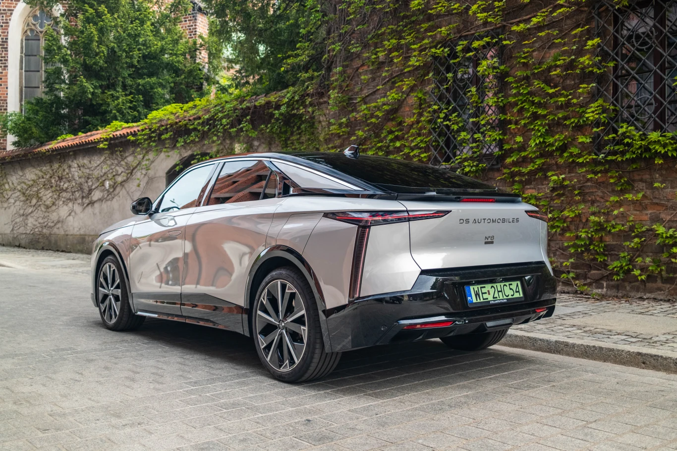 Srebrny nowoczesny samochód elektryczny o eleganckiej sylwetce i czarnym dachu zaparkowany przy brukowanej ulicy, ściana porośnięta bluszczem w tle.