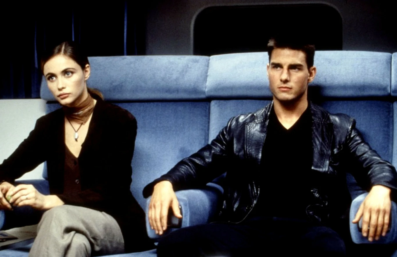 Emmanuelle Beart i Tom Cruise w filmie "Mission: Impossible" Dwie osoby siedzące obok siebie na jasnoniebieskich fotelach, jedna ubrana w czarną skórzaną kurtkę, druga w brązową koszulę i szal, obie patrzące poważnie przed siebie.