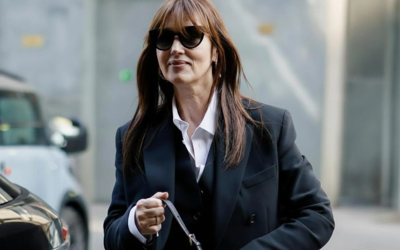 Monica Bellucci Monica Bellucci w ciemnej marynarce i białej koszuli, z brązowymi włosami i okularami słonecznymi, trzyma pasek lub torebkę.