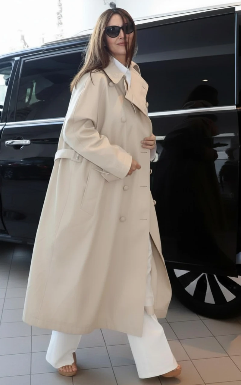 Monica Bellucci Kobieta w długim, beżowym trenczu, białych spodniach i okularach przeciwsłonecznych idzie obok czarnego samochodu.