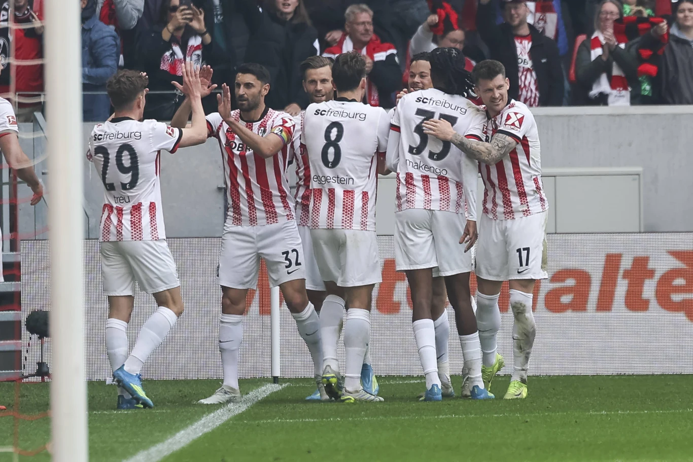 Grupa piłkarzy drużyny w biało-czerwonych koszulkach świętuje zdobycie bramki, stojąc razem na murawie stadionu i okazując radość gestami i przytuleniami.