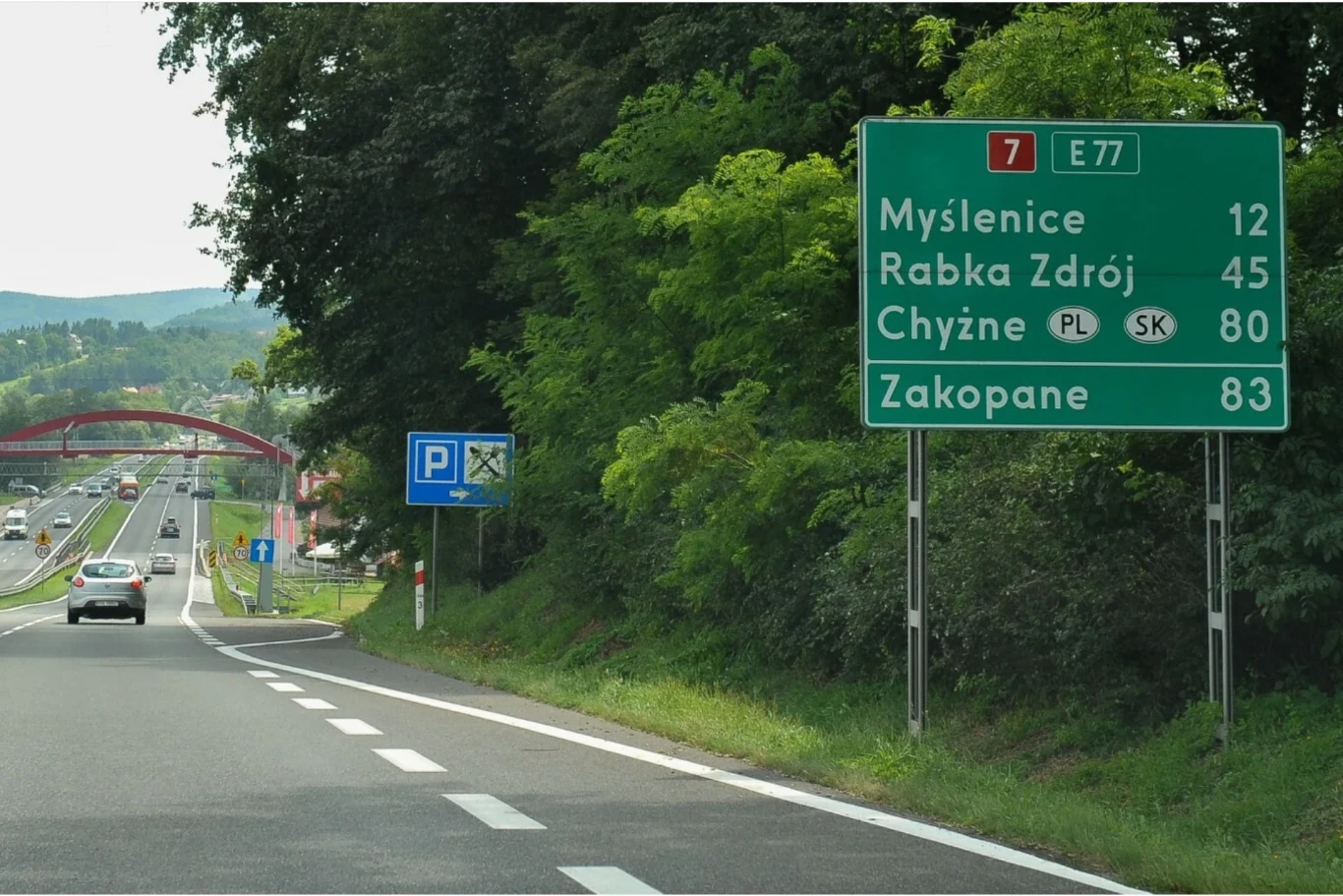 Zielona tablica drogowa z kierunkami do Myślenic, Rabki Zdrój, Chyżnego i Zakopanego przy dwupasmowej drodze, obok znak informujący o parkingu, w tle wiadukt i jadące samochody.