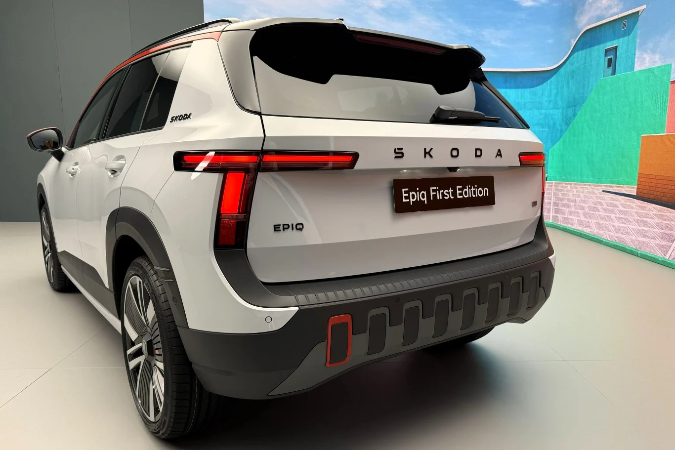 Biały SUV marki Skoda z czarnym dachem, charakterystycznym poziomym pasem świateł z tyłu oraz napisem „Epiq First Edition” na klapie bagażnika, ustawiony na tle kolorowej, geometrycznej ściany.
