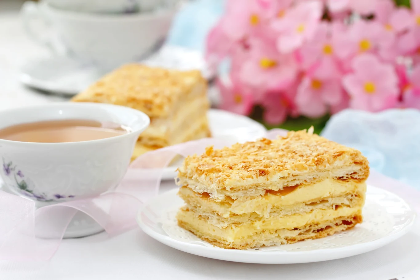 Kapryśna dama to pyszne i proste ciasto bez pieczenia Porcja ciasta Kapryśna Dama z warstwami herbatników i kremu budyniowego na talerzu, z herbatą i kwiatami w tle.