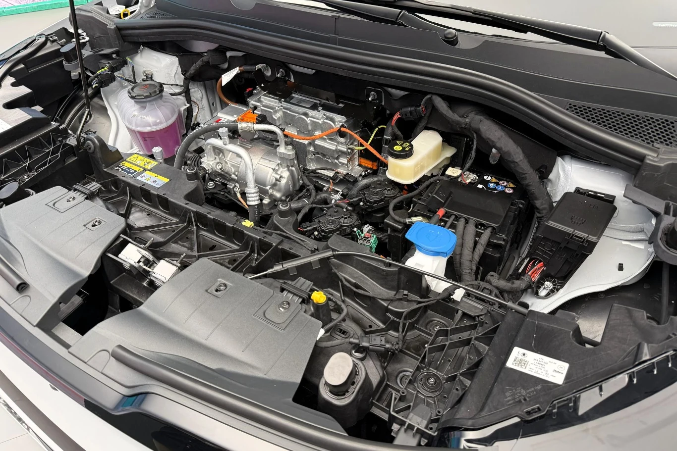 Przestrzeń komory silnika samochodu hybrydowego z widocznym silnikiem elektrycznym, elementami wysokiego napięcia oznaczonymi pomarańczowymi przewodami oraz podzespołami układu chłodzenia i płynów eksploatacyjnych.