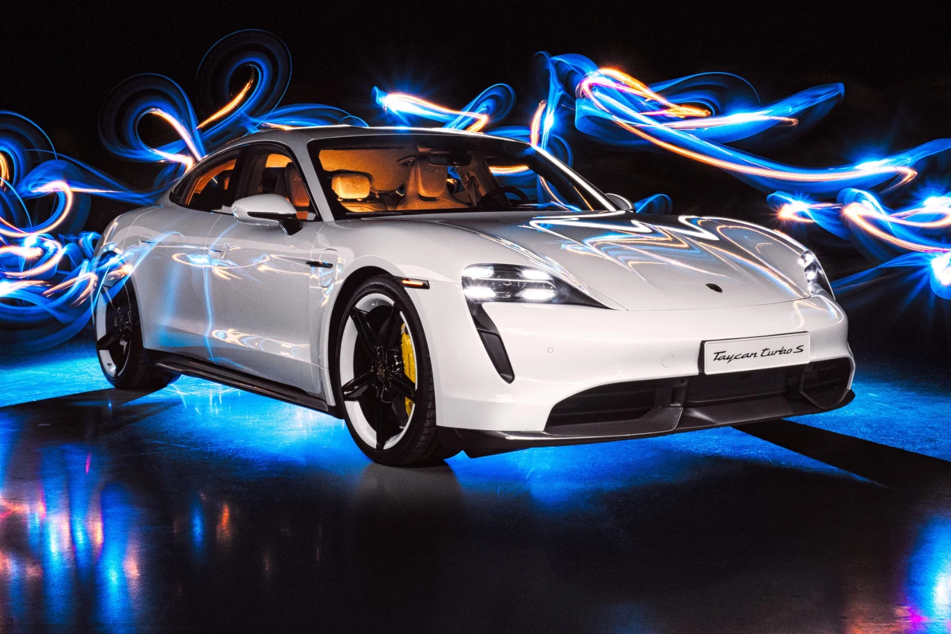 Biały sportowy samochód Porsche Taycan Turbo S na lśniącym podłożu, otoczony dynamicznymi, niebiesko-pomarańczowymi smugami światła na tle ciemności.