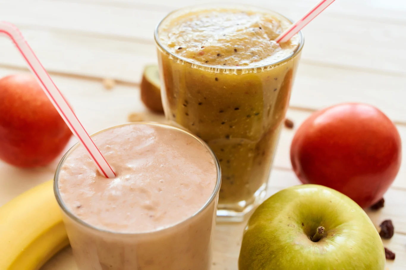 Dwa szklane kubki wypełnione gęstymi smoothie z kolorowymi słomkami, otoczone świeżymi owocami: bananem, zielonym jabłkiem i czerwoną jabłką.