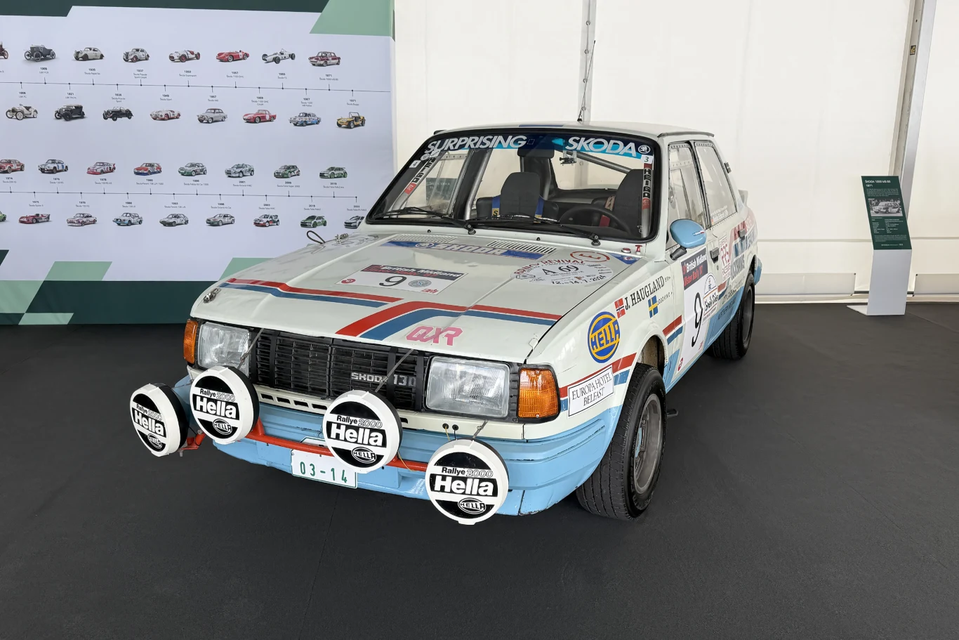 Skoda Favorit w rajdowym malowaniu, wyposażona w dodatkowe lampy na przednim zderzaku oraz liczne naklejki sponsorskie na karoserii, ustawiona na czarnym podłożu w przestronnej, jasnej hali.